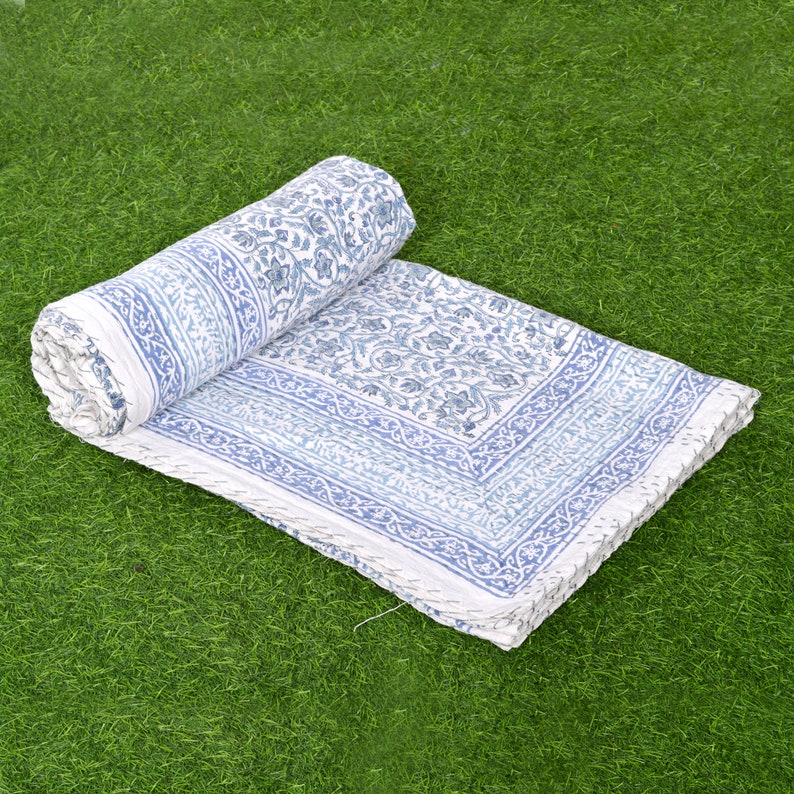 May include: A white cotton quilt with a blue floral pattern. The quilt is rolled up and lying on a green grassy surface.