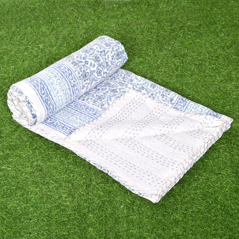 May include: A white and blue kantha quilt with a floral pattern. The quilt is folded and rolled up, and the fabric is soft and textured.