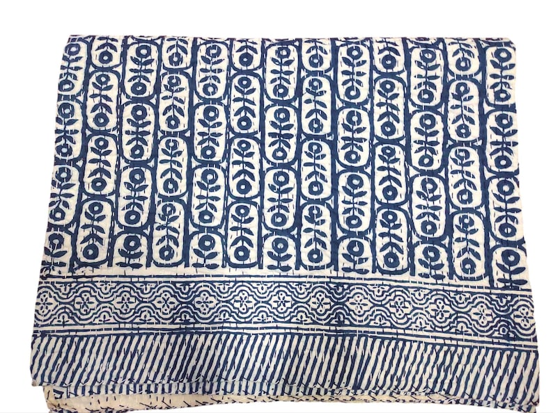K&ouml;nnte beinhalten: Eine wei&szlig;e Baumwolldecke mit einem blauen und wei&szlig;en geometrischen Muster. Die Decke hat einen Rand mit einem sich wiederholenden Muster aus Blumen und Bl&auml;ttern.