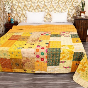 May include: A patchwork quilt in shades of yellow, orange, and cream covers a bed. The quilt features a variety of patterned squares, including floral and geometric designs. The bed is made with white pillows and a gold patterned wallpaper.