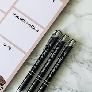 May include: Three black pens with silver accents on a white marble surface. Each pen has the text "Everyone deserves a voice" engraved on the side.