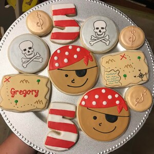 May include: A collection of decorated sugar cookies on a silver platter. The cookies feature pirate faces, skull and crossbones, treasure maps, dollar signs, and the number 5. The name "Gregory" is written on one of the treasure map cookies.