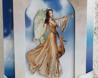 May include: A Christmas card featuring a gold angel with a sparkling halo and wings, holding a trumpet. The angel is standing in a blue sky with clouds. The card has a white border and the words "Merry Christmas" are written in a scroll at the top.