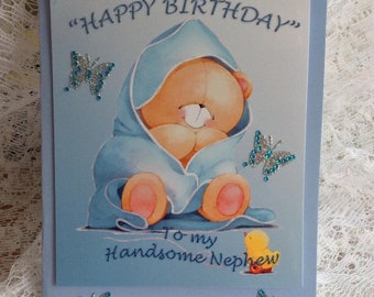 May include: A light blue birthday card with the text "HAPPY BIRTHDAY" and "To my Handsome Nephew". The card features an illustration of a teddy bear in a blue blanket and three glittery butterfly embellishments. A small yellow duck is also present.
