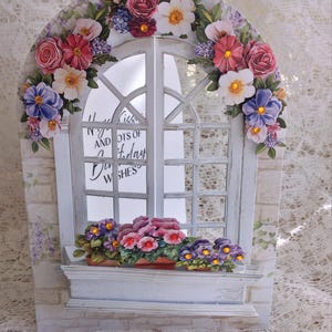 May include: A decorative greeting card shaped like a white window, adorned with colorful paper flowers. The window frame is surrounded by pink, red, and purple flowers. The card has the text "Happy Birthday Wishes".