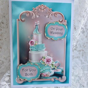 May include: A turquoise and white wedding card with a decorative frame and a cake with a couple on top. The card says "On Your Wedding" and "For You Both".