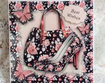 Op de afbeelding: Een wenskaart met een zwarte handtas en een high-heeled schoen, beide versierd met een bloemenpatroon van roze rozen. Roze vlinders en de woorden "Best Wishes" zijn ook aanwezig. De kaart heeft een decoratieve bloemenrand.
