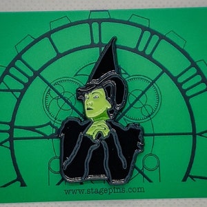 Puede incluir: Un pin de esmalte verde y negro con un personaje del musical Wicked. El personaje lleva un sombrero y un vestido negros y tiene la piel verde. El pin está sobre un fondo verde con un diseño de esfera de reloj.