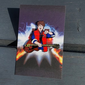 May include: An enamel pin featuring a cartoon character playing a guitar. The character is wearing a red jacket and a blue shirt. The guitar is black with a yellow pickguard. The background is a blurry image of a city street.