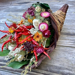 May include: A woven cornucopia overflowing with a colorful arrangement of dried flowers, red chili peppers, and decorative elements. The horn of plenty is made of woven brown material and sits on a wooden surface.