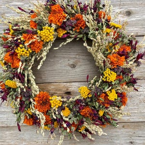May include: A circular wreath composed of dried flowers and foliage in vibrant shades of orange, deep purple, and bright yellow. The arrangement features clusters of marigolds, small yellow button flowers, and dried wheat stalks arranged in a ring.