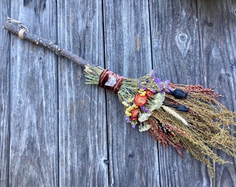 Puede incluir: Un mango de escoba de madera rústica con un manojo de flores secas y hierbas atadas con una cinta marrón. Las flores incluyen flores moradas, amarillas, naranjas y blancas. La escoba se apoya en una pared de madera desgastada.