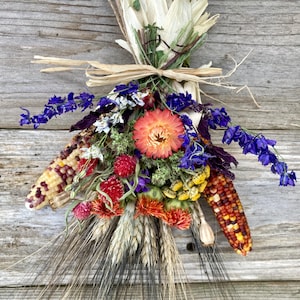 May include: A dried flower arrangement with purple, blue, and yellow flowers, wheat stalks, and colorful corn cobs tied together with twine. The arrangement is hanging on a rustic wooden background.