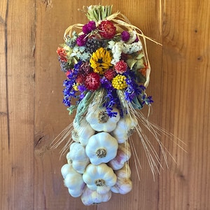 May include: A decorative garlic braid with a colorful arrangement of dried flowers. The flowers include yellow, red, purple, and blue blooms. The garlic bulbs are white and arranged in a cascading pattern. The braid is tied with natural twine and wheat stalks.