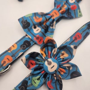 May include: A blue dog collar with a guitar print and a matching bow tie and flower. The bow tie is a classic bow shape and the flower is a five-petal flower.
