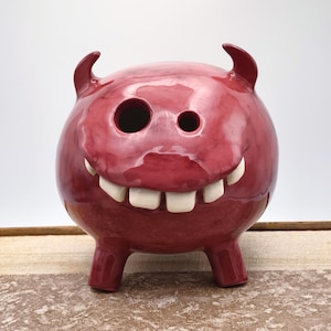 Op de afbeelding: Een glanzend, rood keramisch spaarvarken in de vorm van een lachende duivel met hoorns en grote witte tanden. Het spaarvarken heeft twee ooggaten en rust op een houten oppervlak.