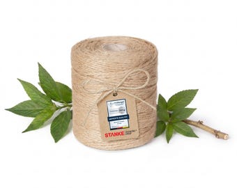 May include: A cylindrical spool of natural tan jute twine featuring a textured, fibrous surface. A brown paper tag is tied to the spool with a piece of the twine, displaying the text "Prüfsiegel", "GEPRÜFTE QUALITÄT", and "STANKE". Green leaves flank the spool.