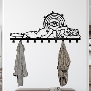 May include: Black metal wall-mounted coat rack featuring a stylized design of a sleeping woman with a celestial eye above her. The rack has 10 hooks and is mounted on a white wall.