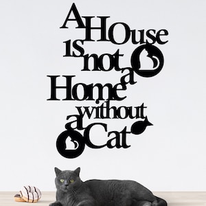 May include: A black and white wall sign with the text "A House is not a Home without a Cat". The sign features a stylised cat silhouette. A grey cat is lying on a wooden table in the foreground.