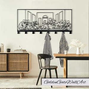 Puede incluir: Arte de pared de metal negro que representa La &Uacute;ltima Cena, con un marco rectangular con una escena de Jes&uacute;s y sus disc&iacute;pulos en una mesa. La obra de arte est&aacute; montada en una pared blanca y tiene ganchos para colgar abrigos u otros art&iacute;culos.
