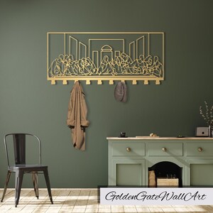 Puede incluir: Arte de pared de metal dorado que representa la &Uacute;ltima Cena con ganchos para colgar abrigos y sombreros. El arte est&aacute; montado en una pared verde con una silla y un gabinete en el fondo.
