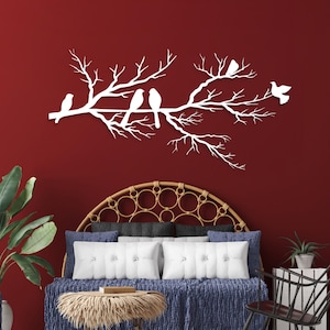 May include: White metal wall art depicting a tree branch with several birds perched on it. The artwork is mounted on a deep red wall. The birds are in various poses, adding a sense of movement to the piece. The overall theme is nature and home decor.