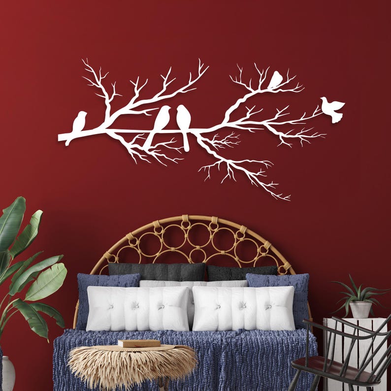May include: White metal wall art depicting a tree branch with several birds perched on it. The artwork is mounted on a deep red wall. The birds are in various poses, adding a sense of movement to the piece. The overall theme is nature and home decor.