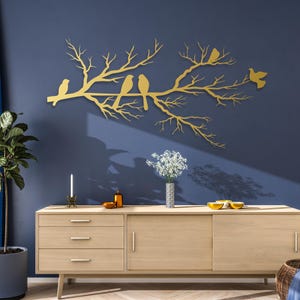 May include: A gold-colored metal wall art piece depicting a tree branch with several birds perched on it. The artwork is mounted on a dark blue wall, creating a striking contrast. The piece adds a touch of nature-inspired decor.