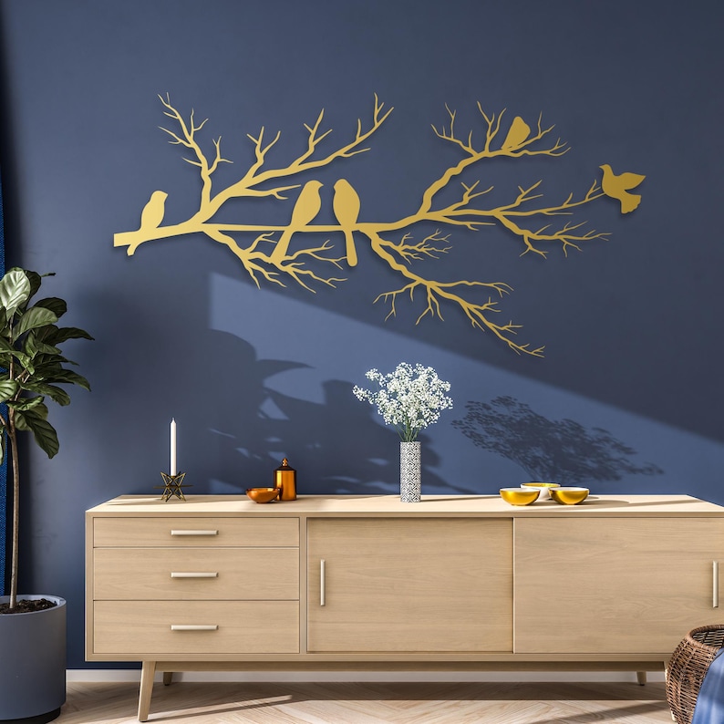 May include: A gold-colored metal wall art piece depicting a tree branch with several birds perched on it. The artwork is mounted on a dark blue wall, creating a striking contrast. The piece adds a touch of nature-inspired decor.