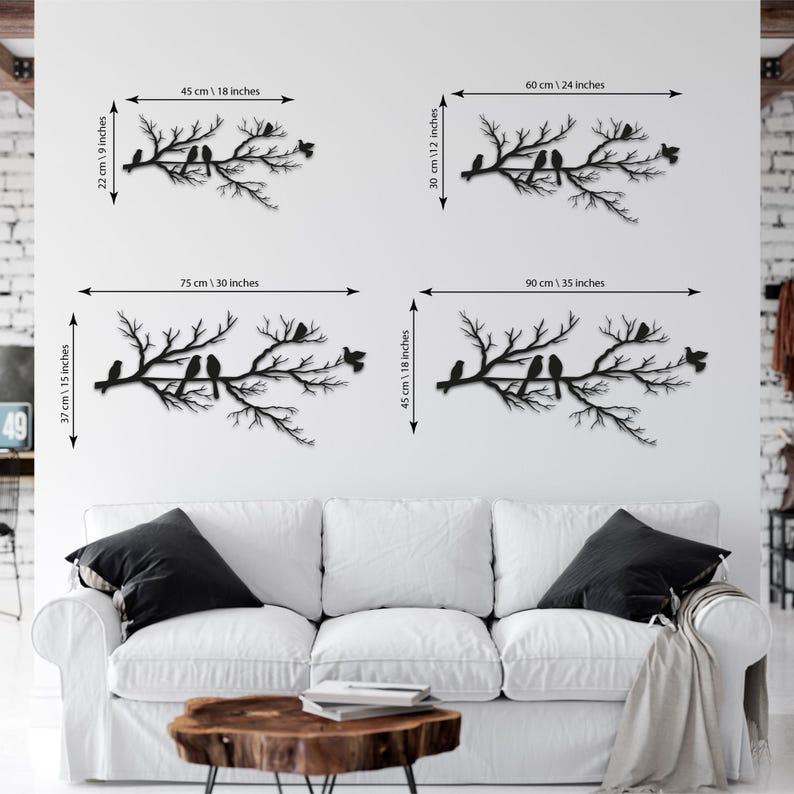 May include: Four black metal wall art pieces depicting branches with birds. The largest piece is 30 inches wide and 15 inches high. The smallest piece is 18 inches wide and 9 inches high. The pieces are displayed above a white sofa.