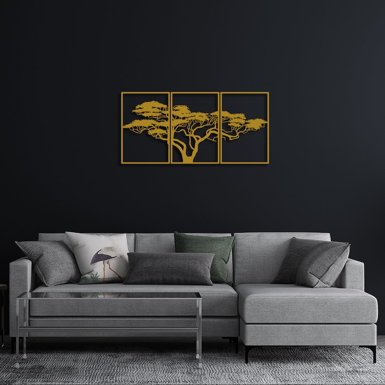 Op de afbeelding: Een gouden metalen wandsculptuur van een boom met takken die zich uitstrekken in drie afzonderlijke frames. De sculptuur hangt aan een zwarte muur boven een grijze hoekbank.