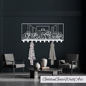 Puede incluir: Arte de pared de metal blanco que representa La &Uacute;ltima Cena, con un perchero con ganchos debajo de la imagen. Dos bufandas cuelgan de los ganchos. El arte est&aacute; montado en una pared gris oscuro con dos sillas y una mesa delante.