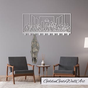 Puede incluir: Perchero de pared de metal blanco con un dise&ntilde;o de la &Uacute;ltima Cena. El perchero tiene varios ganchos y est&aacute; colgado en una pared gris. Dos sillas y una mesa auxiliar est&aacute;n en primer plano.