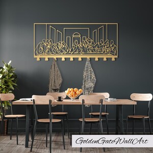 Puede incluir: Una obra de arte de pared de metal dorado que representa la &Uacute;ltima Cena con ganchos para colgar objetos. La obra est&aacute; montada en una pared gris oscuro sobre una mesa de comedor de madera con sillas de metal negro.