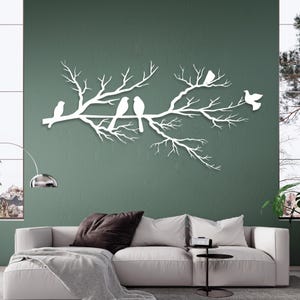 May include: White metal wall art depicting a tree branch with several birds perched on it. The birds and branches are cut out in a minimalist style, creating a clean and modern aesthetic. The artwork is mounted on a green wall.