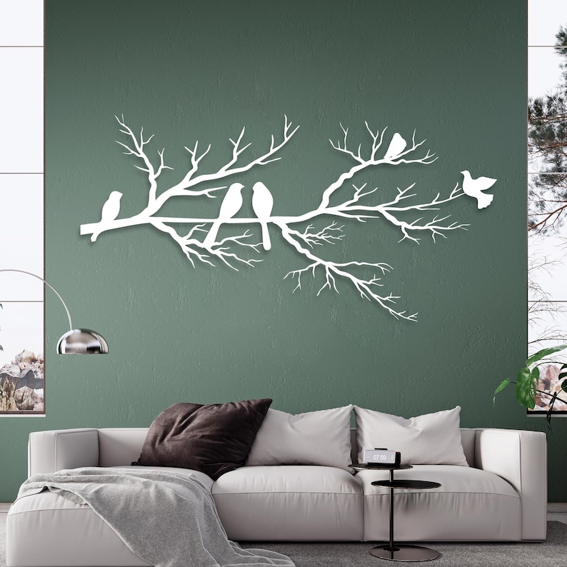 May include: White metal wall art depicting a tree branch with several birds perched on it. The birds and branches are cut out in a minimalist style, creating a clean and modern aesthetic. The artwork is mounted on a green wall.