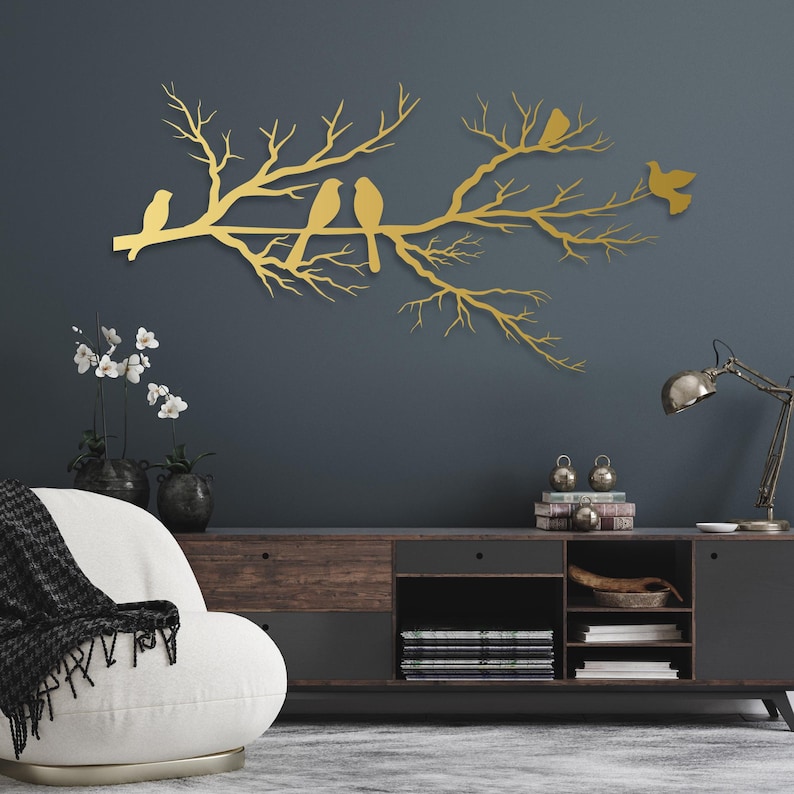 May include: A gold-colored metal wall art piece depicting a tree branch with several birds perched on it. The birds and branches are in a silhouette style, creating a decorative and artistic element for home decor.