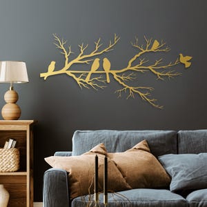 May include: A gold-colored metal wall art piece depicting a tree branch with several birds perched on it. The artwork is mounted on a dark gray wall above a blue sofa with brown pillows. The birds are stylized and add a touch of nature to the decor.