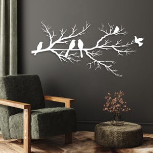 May include: A white metal wall art piece depicting a tree branch with several birds perched on it. The birds are stylized and the branch has many small twigs. The art is mounted on a dark gray wall, creating a contrast.