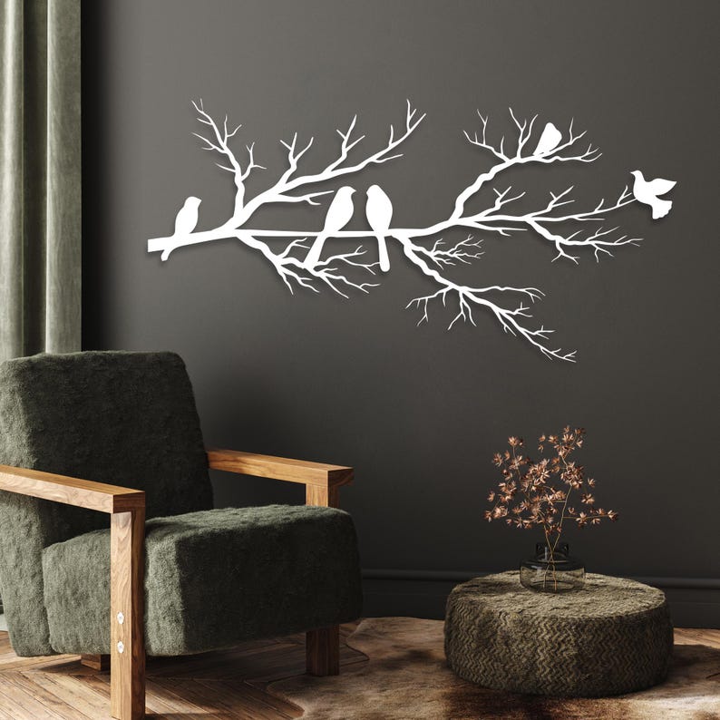 May include: A white metal wall art piece depicting a tree branch with several birds perched on it. The birds are stylized and the branch has many small twigs. The art is mounted on a dark gray wall, creating a contrast.