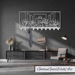 Puede incluir: Arte de pared de metal blanco que representa La &Uacute;ltima Cena con un perchero debajo. El arte est&aacute; montado en una pared gris con una c&oacute;moda de madera y una silla en primer plano.
