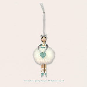 May include: A hanging ornament featuring a wooden peg doll with a painted face, brown hair, and a silver bow. The doll wears a white fluffy pom-pom skirt, a silver ribbon necklace with a teal glitter heart, and teal painted ballet slippers.