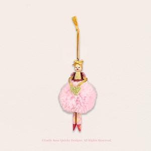 May include: A hanging ornament featuring a small wooden figure with a painted face and blonde hair, wearing a dark pink bodice and a large, fluffy light pink pom-pom skirt. The figure holds a gold glitter heart and has painted pink legs.
