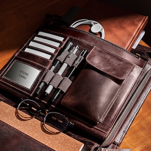 May include: A brown leather organizer with multiple compartments, including slots for cards and pens. The organizer contains pens, a notebook, glasses, and a store card. A pair of white wireless earbuds and a brown notebook are also visible.