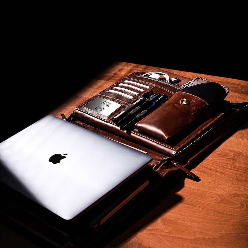 Puede incluir: Un malet&iacute;n de cuero marr&oacute;n con un port&aacute;til plateado, bol&iacute;grafos y un estuche para gafas. El malet&iacute;n tiene m&uacute;ltiples compartimentos, incluyendo ranuras para tarjetas y un portal&aacute;pices. El texto "STORE CARD" es visible.