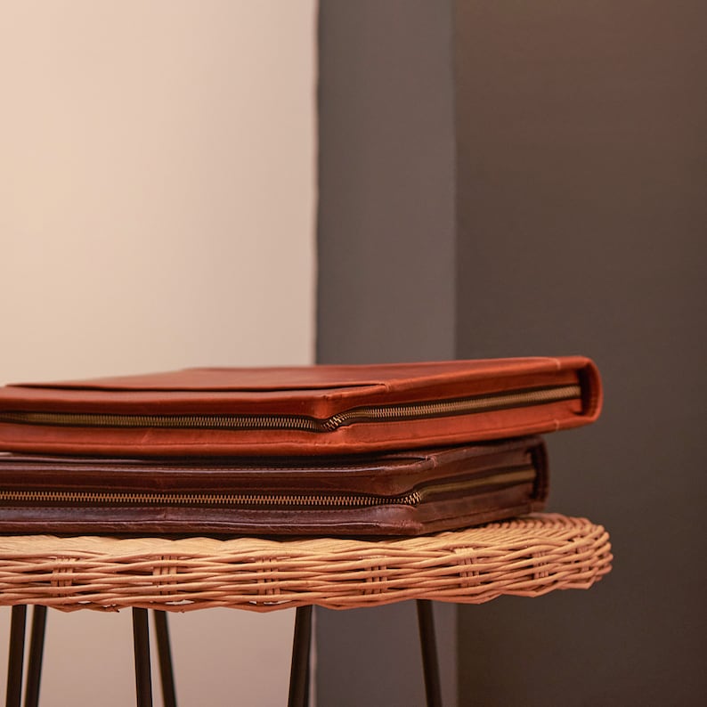Puede incluir: Dos fundas de documentos de cuero apiladas sobre una mesa de mimbre. La funda superior es de color marr&oacute;n c&aacute;lido, la inferior es de color marr&oacute;n m&aacute;s oscuro. Ambas tienen un cierre de cremallera color lat&oacute;n. La mesa tiene patas de metal negro.
