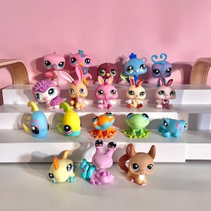 Puede incluir: Una colección de pequeñas figuras de animales coloridos, incluyendo pájaros, conejos, ranas y un caracol. Las figuras son de varios colores como rosa, azul, verde y amarillo, con ojos grandes y expresivos. Están dispuestas en una estantería blanca sobre un fondo rosa.