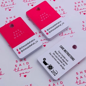 K&ouml;nnte beinhalten: Rosa quadratische H&auml;ngeetiketten mit dem Text "She Said What?" und Social-Media-Handles. Wei&szlig;e quadratische H&auml;ngeetiketten mit dem Text "Pflegehinweise" und Waschanweisungen.