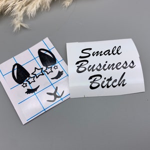 May include: Two white stickers, one with a black cartoon face with eyelashes and stars, and the other with the text "Small Business Bitch" in black cursive.
