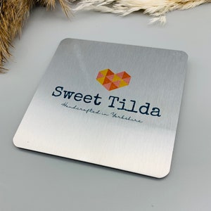 May include: A silver metal sign with a heart-shaped logo in shades of orange, yellow, and pink. The text "Sweet Tilda" is printed in black with the tagline "Handcrafted in Yorkshire" below.